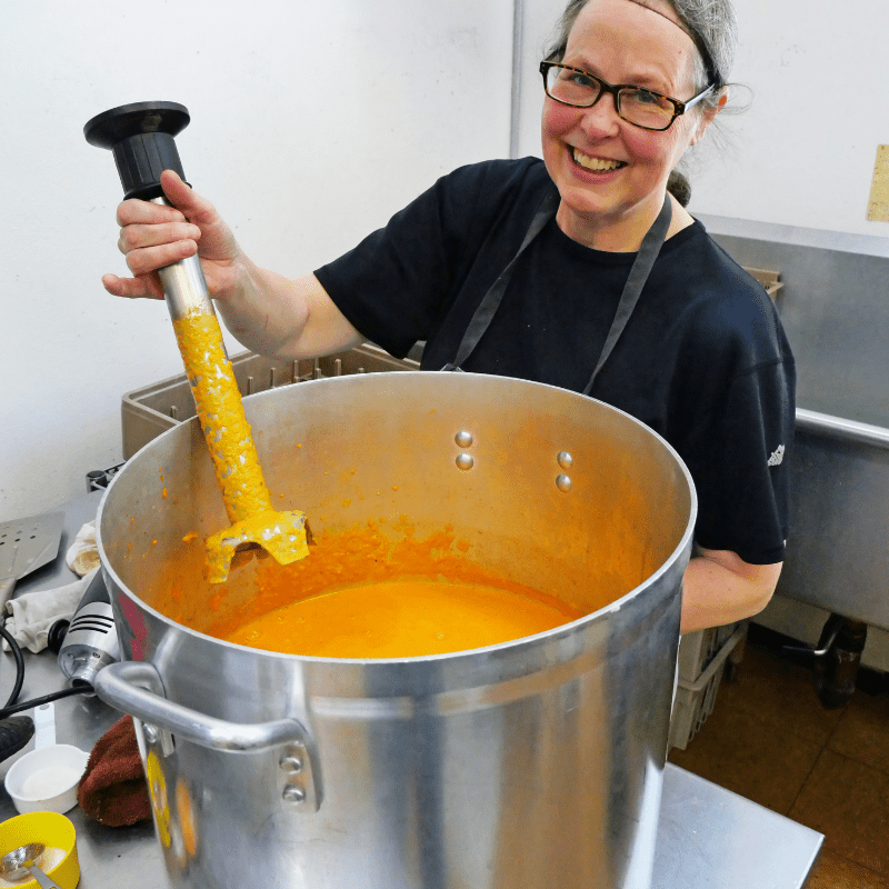 Smiling women holds large immersion blender over giant pot of carrot soup