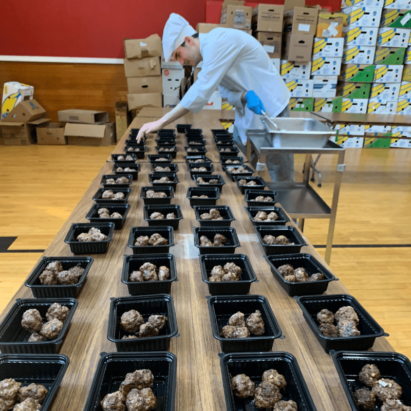 Man stands over table portioning meatballs into rows of containers