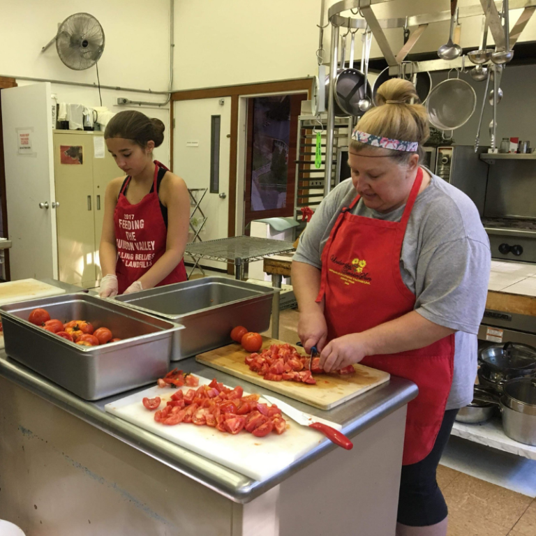 A woman and teenage girl wear red aprons while chopping tomatoes