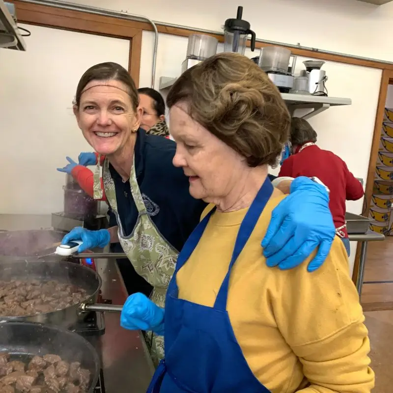 two women in aprons cooking at a stove, one smiles with her arm around the other