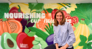 painted mural of assorted vegetables with words "nourishing our neighbors" slightly blurry background with a woman in front smiling at the camera.