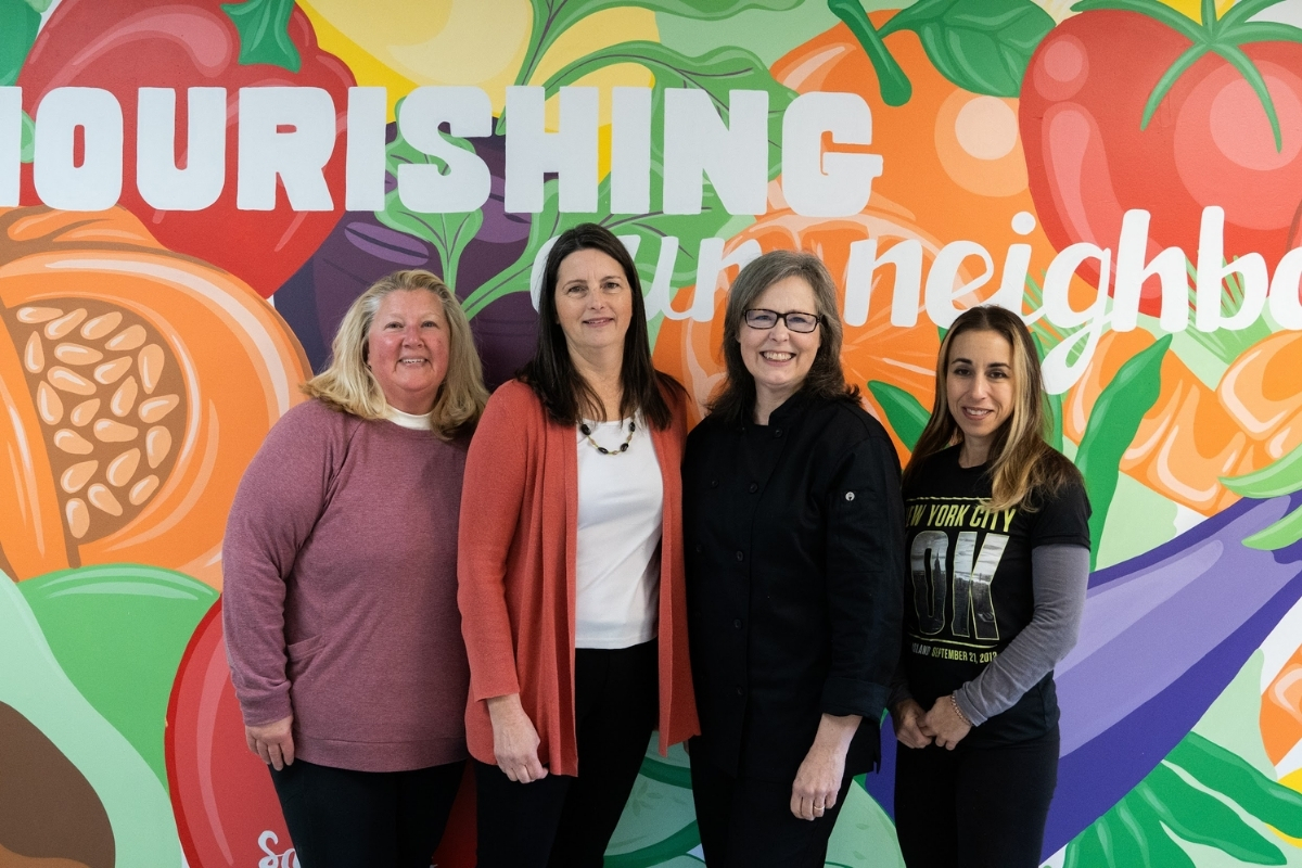 Four women stand in front of a colorful mural with the painted words "Nourishing Our Neighbors"