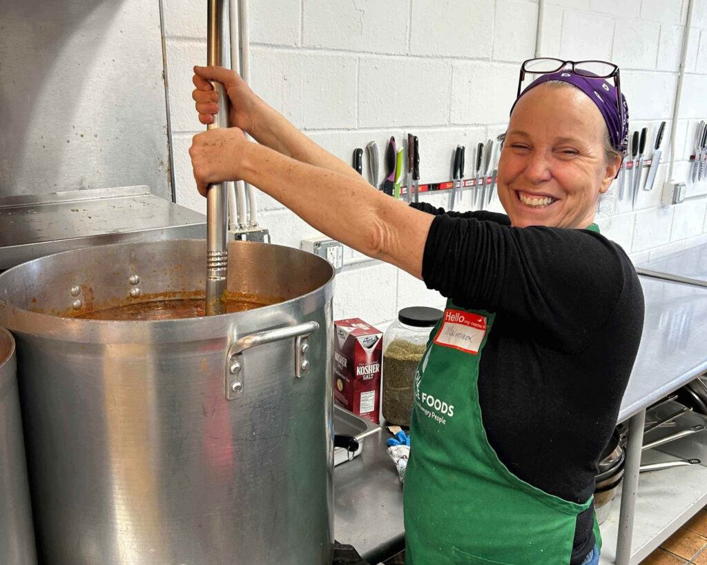 Woman in Second Chance Foods apron stirs a very large pot filled with soup