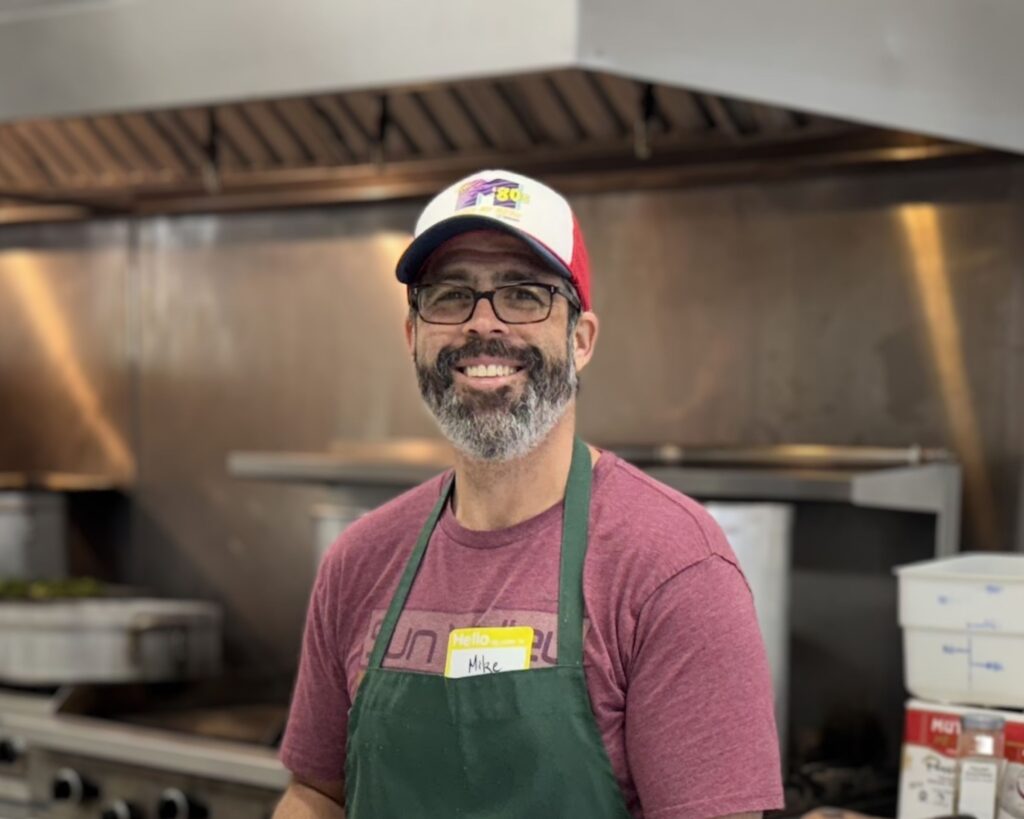 man in green apron and ball cap smiles in a front of an industrial kitchen stove and hood