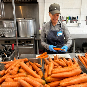A woman peeling a carrot with large bins of unpeeled carrots in front of her